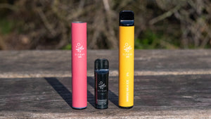 Three vape pens in pink, black, and yellow on a wooden surface.